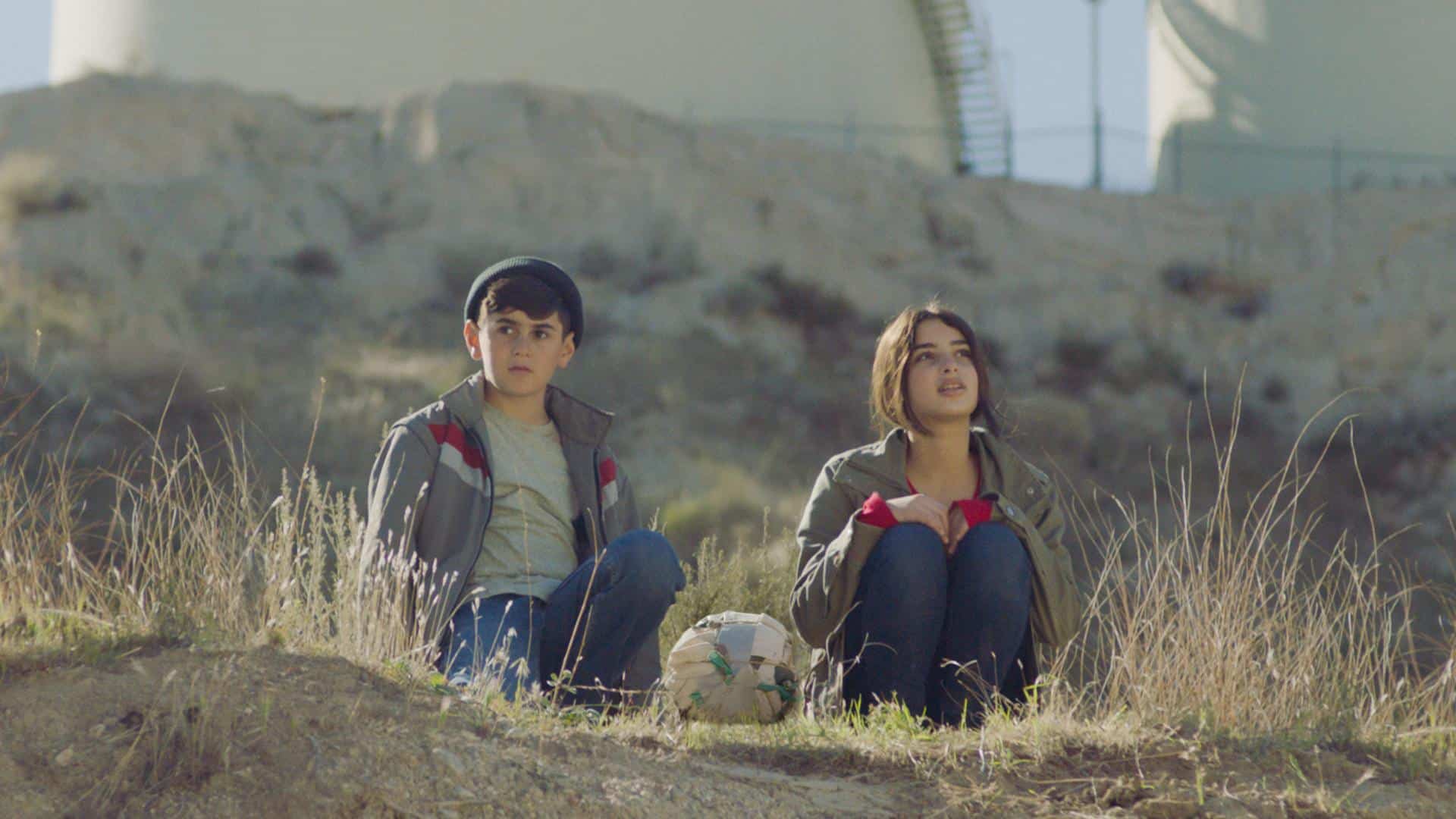 A film still from Bukra shows a young Syrian boy and girl sitting in a field. Both of their gazes in the distance. Between them is a makeshift soccer ball made with different fabrics, held together with bands.