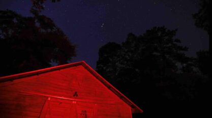 A film still from Celestine Florida Storm shows a barn illuminated in red light in the middle of the night. The shadows of trees can be seen above the barn against the starry sky.