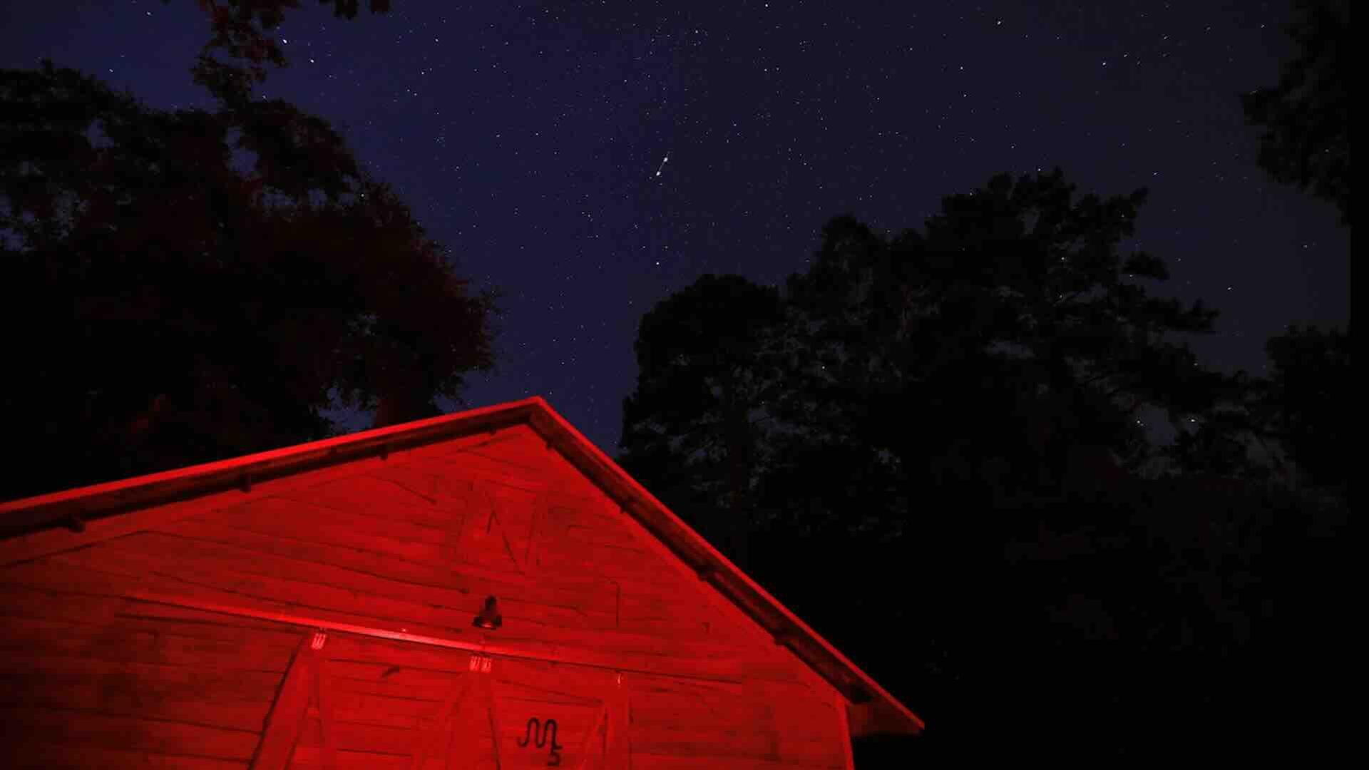 A film still from Celestine Florida Storm shows a barn illuminated in red light in the middle of the night. The shadows of trees can be seen above the barn against the starry sky.