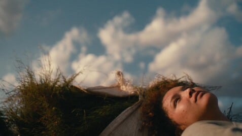 A film still from Carissa shows a young girl lying against white bags filled with a green crop. The girl is looking into the partly cloudy blue sky.