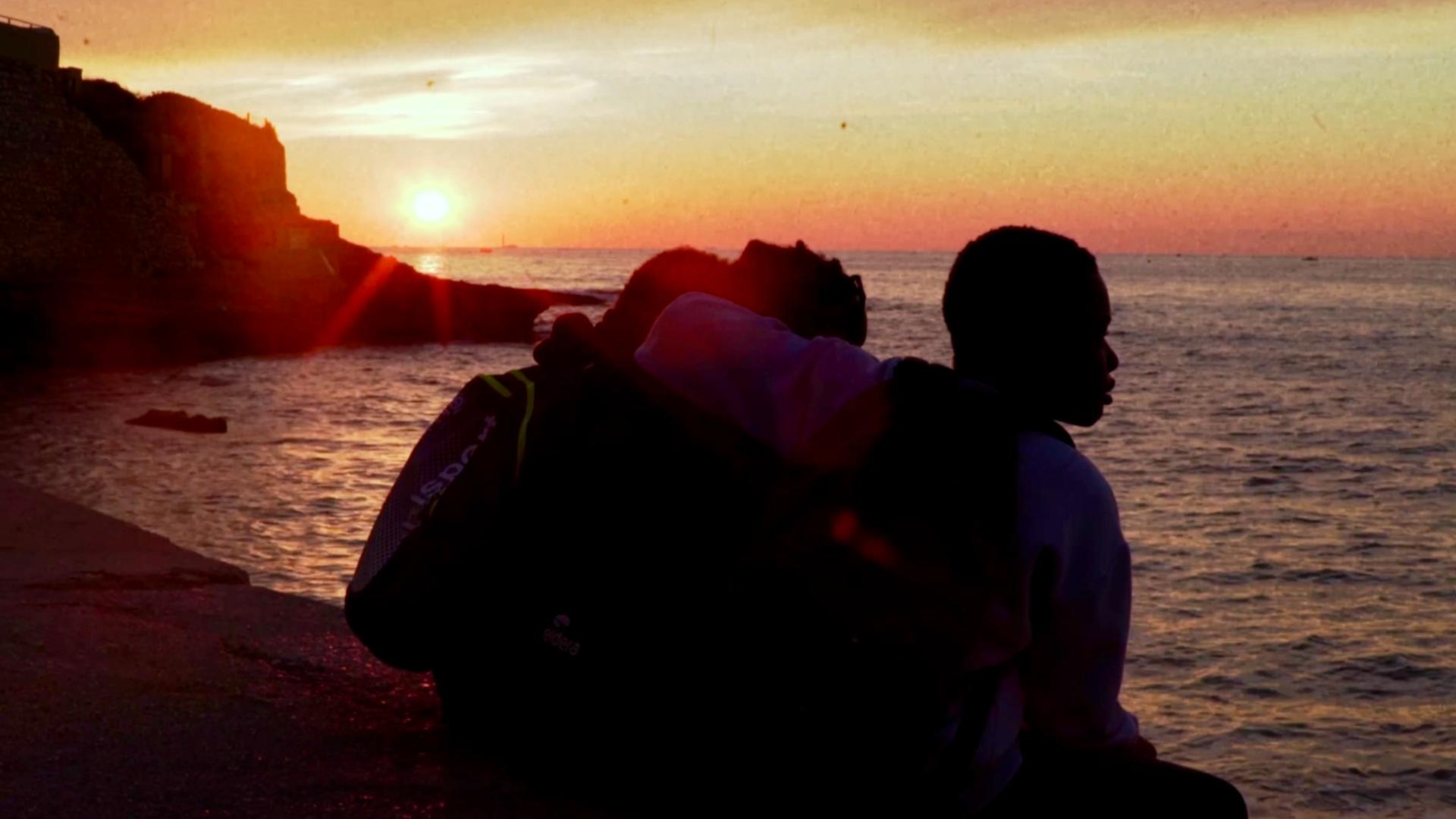 A poster for Children of the Waves features three film stills stacked vertically, each depicting the same three young Black boys. In the top still, they sit on a ledge in front of a white building with blue shutters, gazing into the distance. The boy on the left holds a Monopoly game in his lap, the middle boy holds a folded piece of paper, and the boy on the right holds a soccer ball. The middle still shows their silhouettes as they sit at the edge of a surface, watching the sunset over the ocean. In the bottom still, they sit in front of the same building from the first still, laughing and talking with each other.