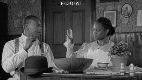 A black-and-white film still from Compensation shows a Black woman and man sitting at a dining table. Both their gazes are fixed on each other as they converse with sign language. Above their heads, the word “F–L–O–W-” appears in small white text with dashes between each letter.