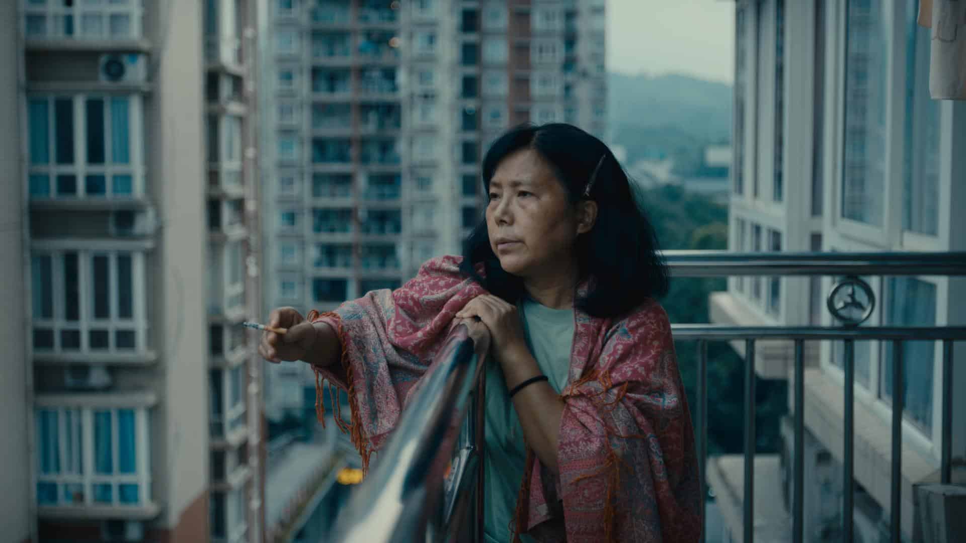 A film still from Correct Me If I’m Wrong shows an older Asian woman standing on a balcony, leaning against the railing as she looks into the distance. One of her arms is perched on the railing with a cigarette in her hand. She wears a pink shawl draped around her shoulders.