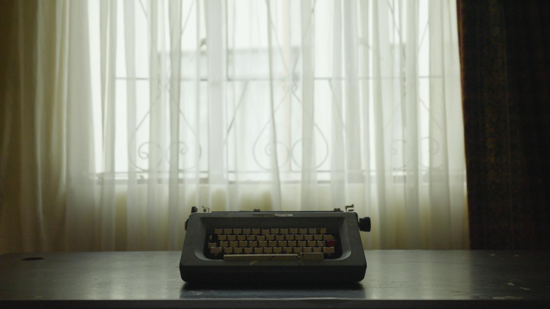 A film still from Dear Sikhonkwane shows a gray typewriter resting on a table in front of a sheer white curtain. Behind the curtain, a window with an iron guard is partially visible.