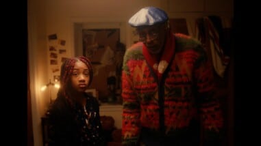 Black actors Damien Wallace and KaCi Camille Ferguson stand next to each other in their character roles, astounded. Damien on the right wears a colorful cardigan and red scarf with a navy blue beret and glasses. KaCi, on his left, wears a polka dot dress and a black cardigan with red box braids. They stand in a low light bedroom