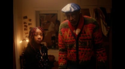 Black actors Damien Wallace and KaCi Camille Ferguson stand next to each other in their character roles, astounded. Damien on the right wears a colorful cardigan and red scarf with a navy blue beret and glasses. KaCi, on his left, wears a polka dot dress and a black cardigan with red box braids. They stand in a low light bedroom