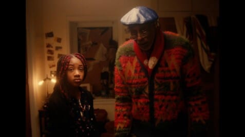 Black actors Damien Wallace and KaCi Camille Ferguson stand next to each other in their character roles, astounded. Damien on the right wears a colorful cardigan and red scarf with a navy blue beret and glasses. KaCi, on his left, wears a polka dot dress and a black cardigan with red box braids. They stand in a low light bedroom