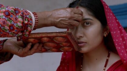 This image captures a culturally rich and intimate moment during a traditional Hindu ritual, likely in Nepal. A young woman, dressed in a vivid red saree with traditional jewelry, sits solemnly as an elder woman applies a red tika (a ceremonial mark) to her forehead. The elder's hand, adorned with red bangles, holds a wooden tray filled with red powder, used in various religious and cultural ceremonies.