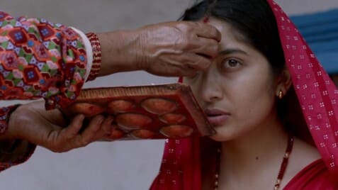 This image captures a culturally rich and intimate moment during a traditional Hindu ritual, likely in Nepal. A young woman, dressed in a vivid red saree with traditional jewelry, sits solemnly as an elder woman applies a red tika (a ceremonial mark) to her forehead. The elder's hand, adorned with red bangles, holds a wooden tray filled with red powder, used in various religious and cultural ceremonies.