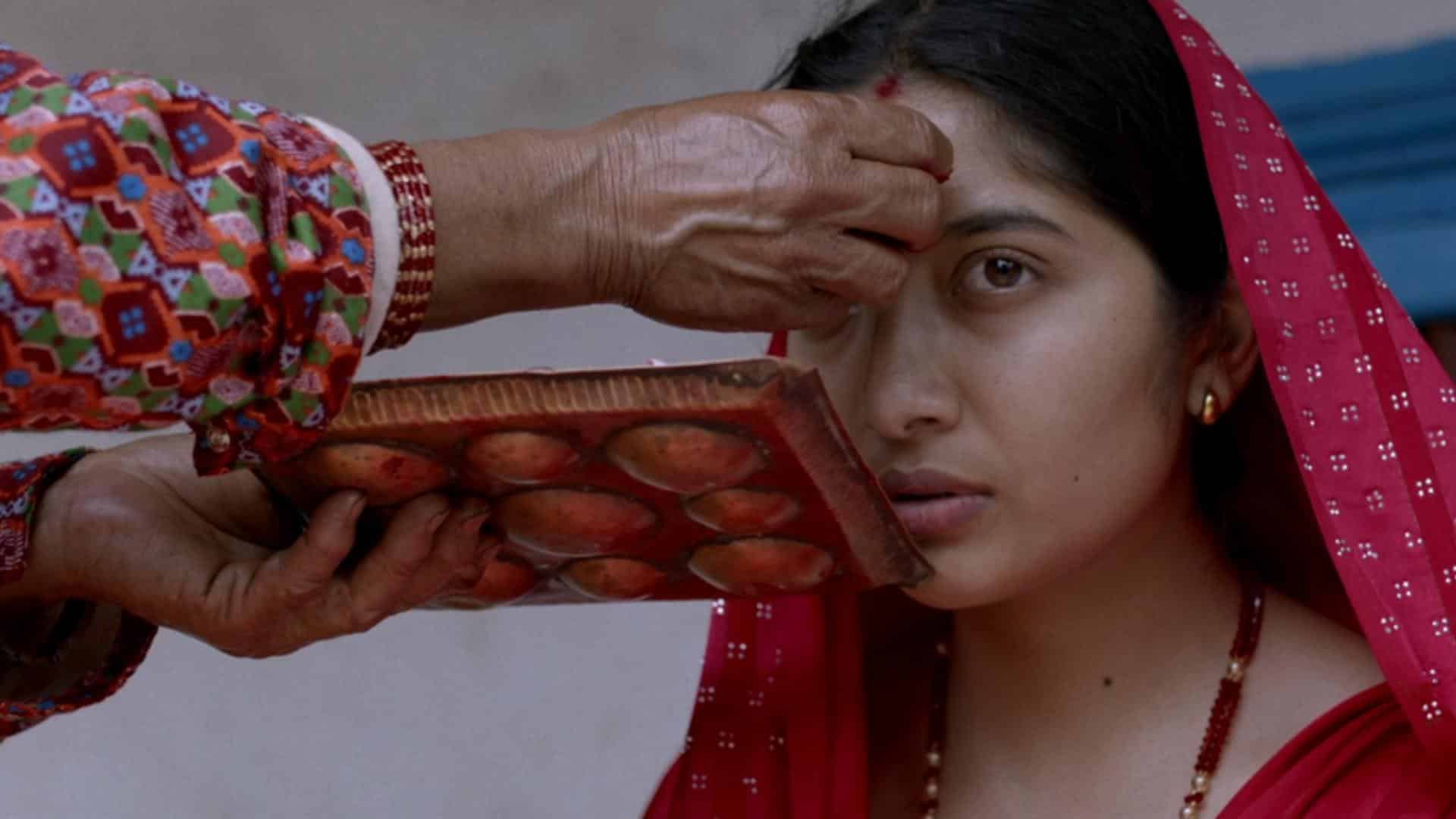 This image captures a culturally rich and intimate moment during a traditional Hindu ritual, likely in Nepal. A young woman, dressed in a vivid red saree with traditional jewelry, sits solemnly as an elder woman applies a red tika (a ceremonial mark) to her forehead. The elder's hand, adorned with red bangles, holds a wooden tray filled with red powder, used in various religious and cultural ceremonies.