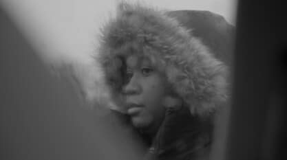 A black-and-white film still from Exodus shows a woman wearing a winter coat with a fur lined hood pulled over her head. The view of the woman is through a gap between two unknown objects.
