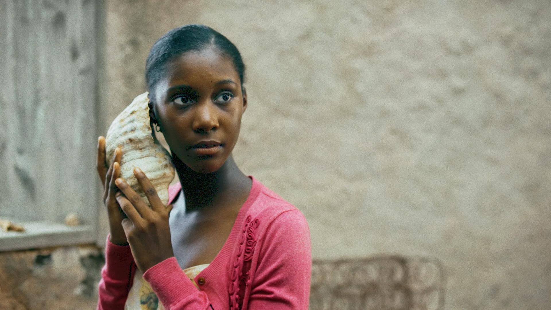 A film still from Hanami shows a young Black girl holding a conch shell up to her ear as she looks at something off screen.
