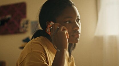 A Black teenage girl sits on her bed in front of a window and talks on an orange cellphone.