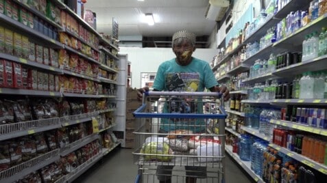 A film still from Hosts for Half a Century shows an older Asian man wearing a face mask pushing a shopping cart filled with groceries down an aisle of a grocery store.