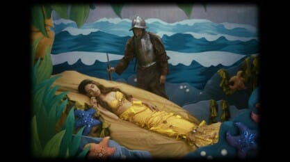 A film still from Las Cosas Que Brillan shows a black frame around a staged scene. Inside the frame is a woman dressed as a mermaid, with a yellow tail and top, sleeping on a faux rock. Standing above her is a man dressed as a soldier, wearing dirty armor with a grimy face, gazing down at her. They are positioned within a theatrical set designed to mimic an ocean scene with a series of waves behind them.