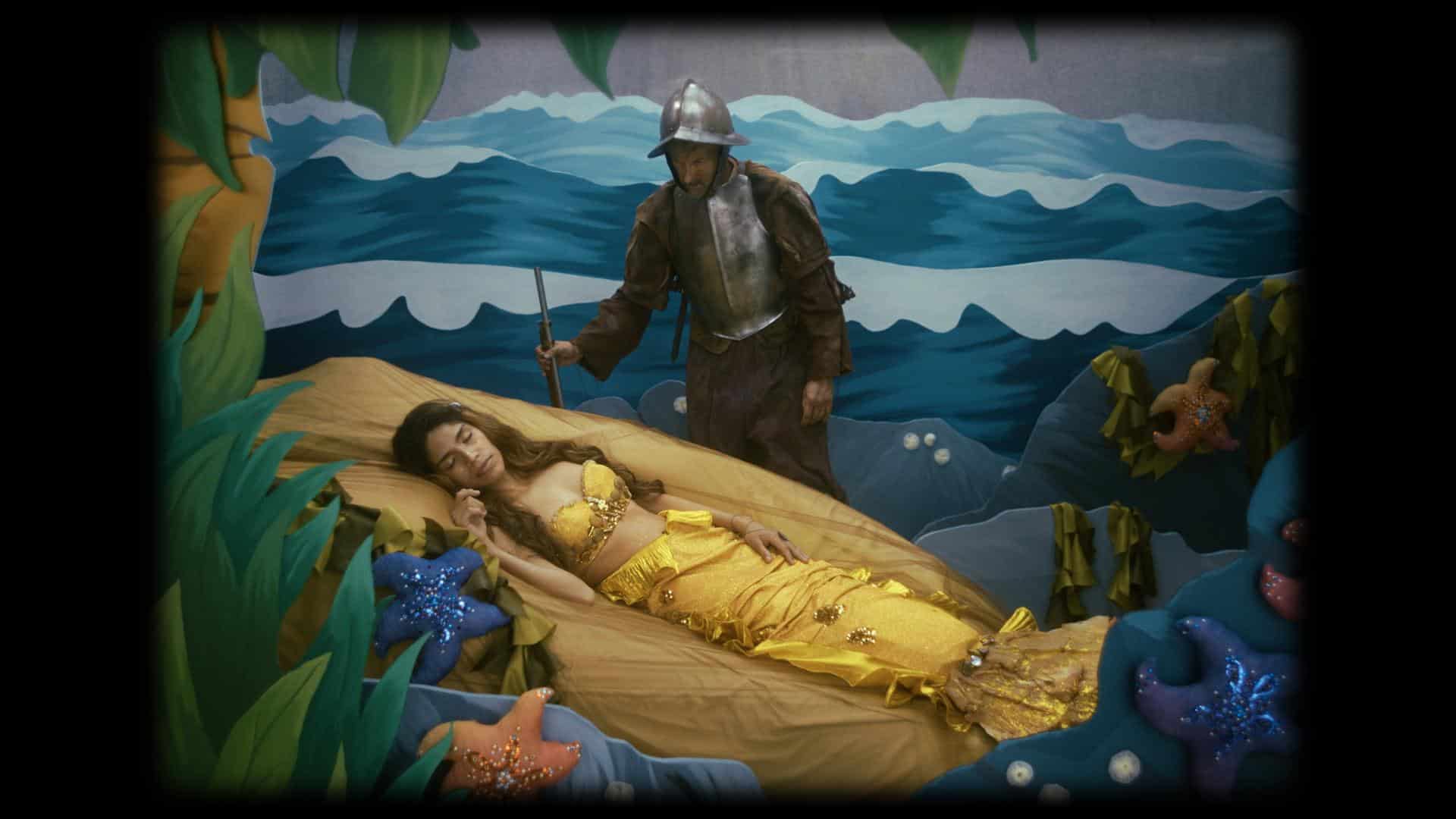 A film still from Las Cosas Que Brillan shows a black frame around a staged scene. Inside the frame is a woman dressed as a mermaid, with a yellow tail and top, sleeping on a faux rock. Standing above her is a man dressed as a soldier, wearing dirty armor with a grimy face, gazing down at her. They are positioned within a theatrical set designed to mimic an ocean scene with a series of waves behind them.