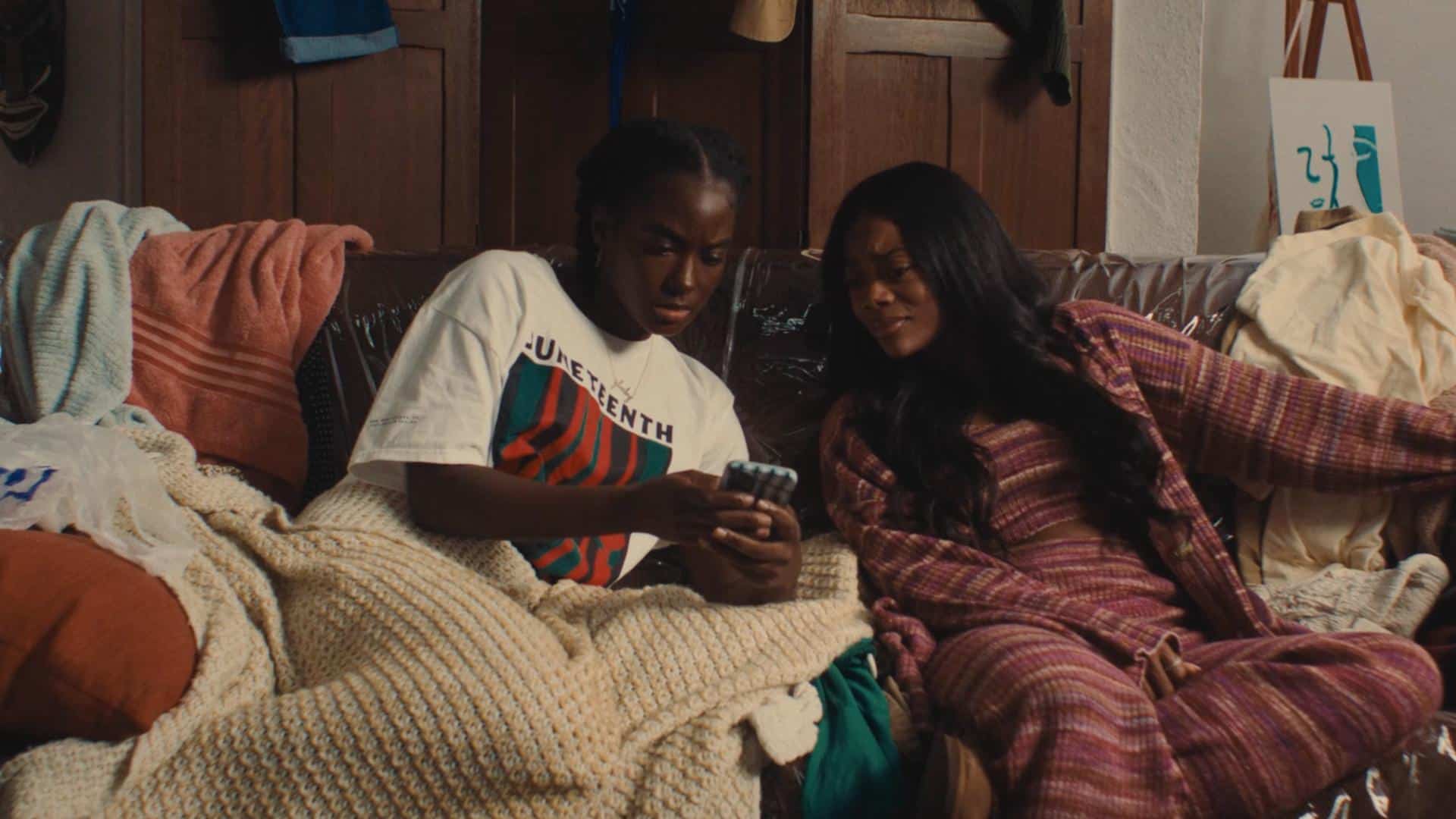 A film still from Last Hoorah at G-Baby’s shows two Black women sitting on a plastic-covered couch. They both look with concern at a phone one of the women holds. They are surrounded by bath towels and other articles of clothing.