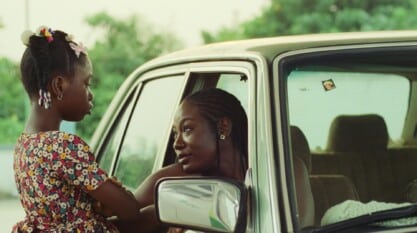 A film still from Leaving Ikorodu in 1999 shows a Black woman looking at a young Black girl, who stands with her arms crossed and a solemn face at her passenger window.