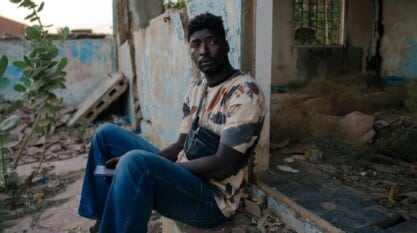 Ousseynou sits quietly in front of an abandoned structure, its walls bare and exposed.