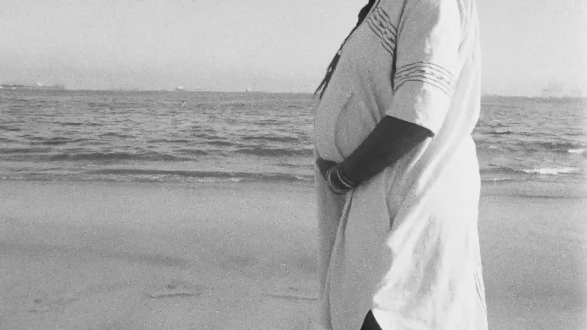 A black-and-white film still from Listen to Me shows presumably a pregnant woman holding her stomach as she walks along the shoreline of a beach.