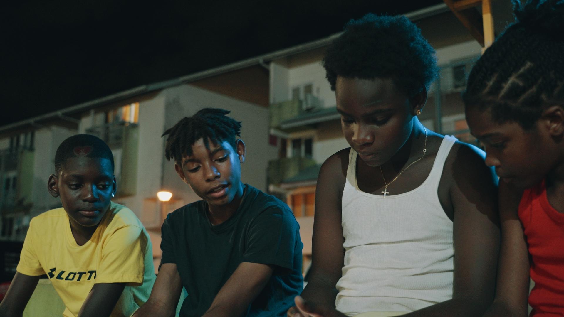 A film still from Listen to the Voices shows four young Black boys sitting side-by-side. Their attention is focused on something that one of the boys holds in his lap off screen.