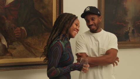 A film still from Love, Brooklyn shows a Black man and woman conversing in front of a wall of framed paintings. They both hold a glass of wine. The man looks at the woman who has her head tilted towards him, smiling with her eyes closed.