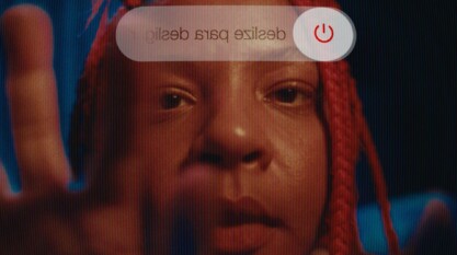 A film still from Lazy White Cows shows the face of a Brazilian girl with red braids from the perspective of her phone. The power off symbol can be seen at the top of the still. Her hand can be seen reaching to turn off her phone.
