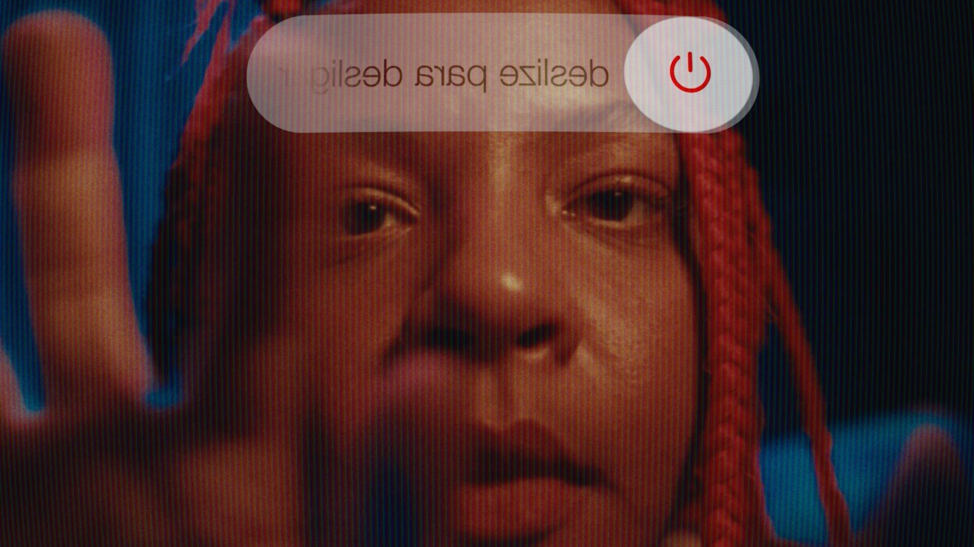 A film still from Lazy White Cows shows the face of a Brazilian girl with red braids from the perspective of her phone. The power off symbol can be seen at the top of the still. Her hand can be seen reaching to turn off her phone.