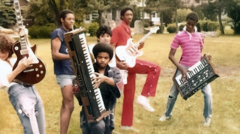 A film still from Move Ya Body: The Birth of House shows a group of six young people. They stand in a yard holding various instruments.