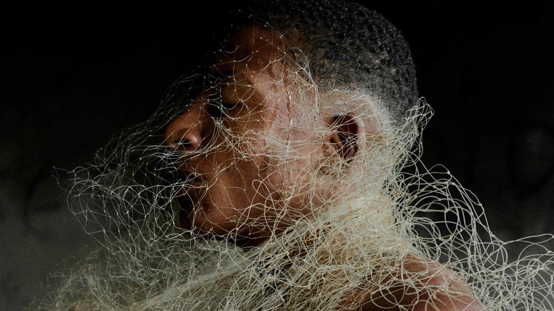 A film still from Natimorto shows the side profile of a Brazilian man. There is white netting that wraps around his head and body.