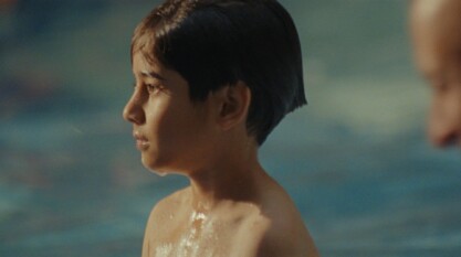 A medium close-up showcasing the profile of a young six-year old boy standing in the middle of a community pool, staring off into the distance, closely fixated on something off-screen.