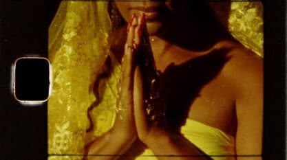 A film still from Oríkì Oshun shows a woman wearing a yellow veil and dress. Her hands, dressed in gold jewelry and red nails, are pressed together in front of her chest.