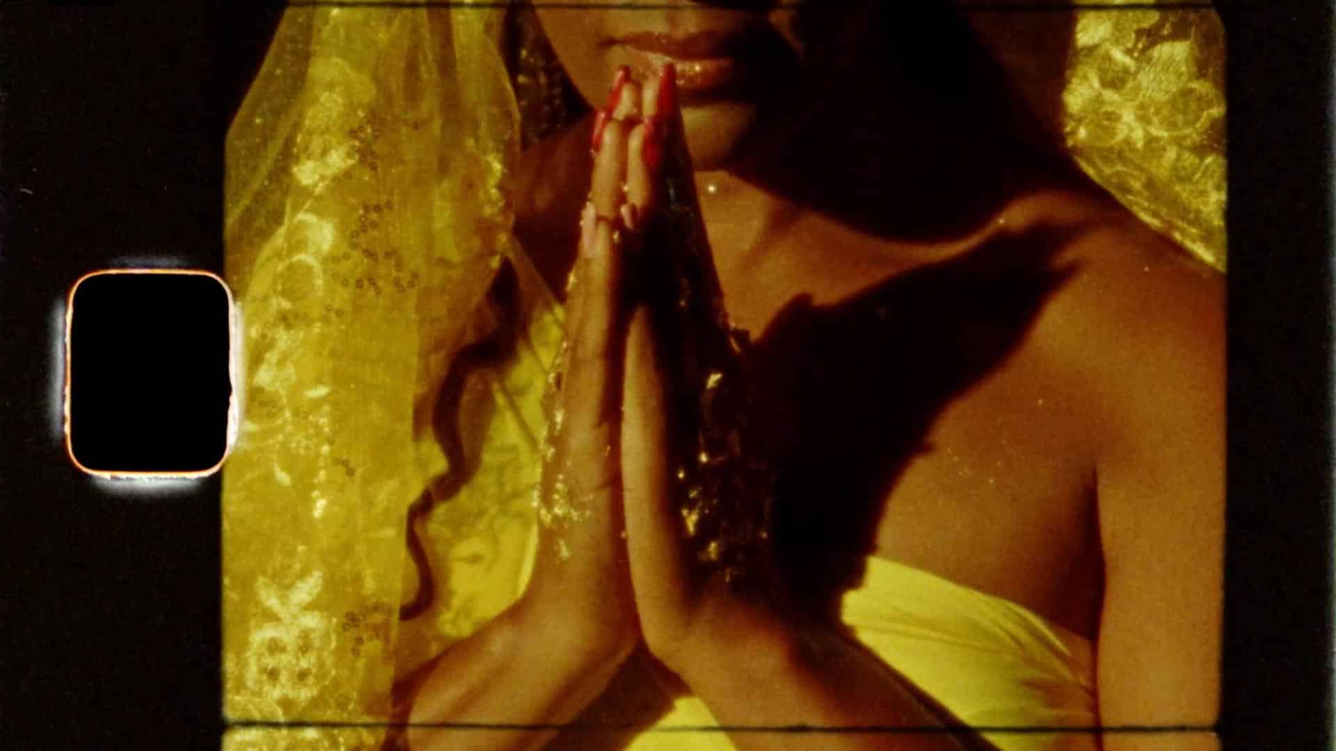 A film still from Oríkì Oshun shows a woman wearing a yellow veil and dress. Her hands, dressed in gold jewelry and red nails, are pressed together in front of her chest.