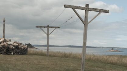 A film still from Otherworld shows a clothesline with empty clothespins standing on a coastline under a cloudy sky. A pile of chopped wood sits in the background.