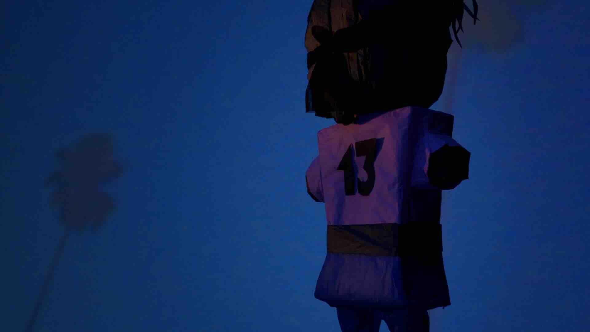 A film still from Piñata Prayers shows the back of a piñata that faces a blue wall with a palm tree pattern. The piñata resembles a person playing a sport. It wears the number thirteen on its back.