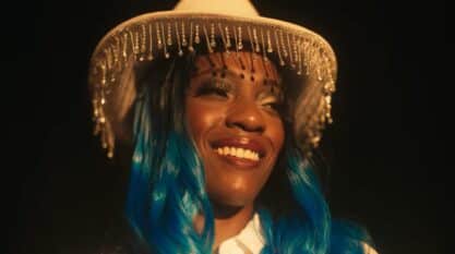 A film still from Ree’s Destiny shows the face of a Black woman who smiles looking into the distance. Her black curled hair fades into a vibrant blue, and she wears a full face of makeup along with a white cowboy hat wrapped with dangling clear crystals around the rim.