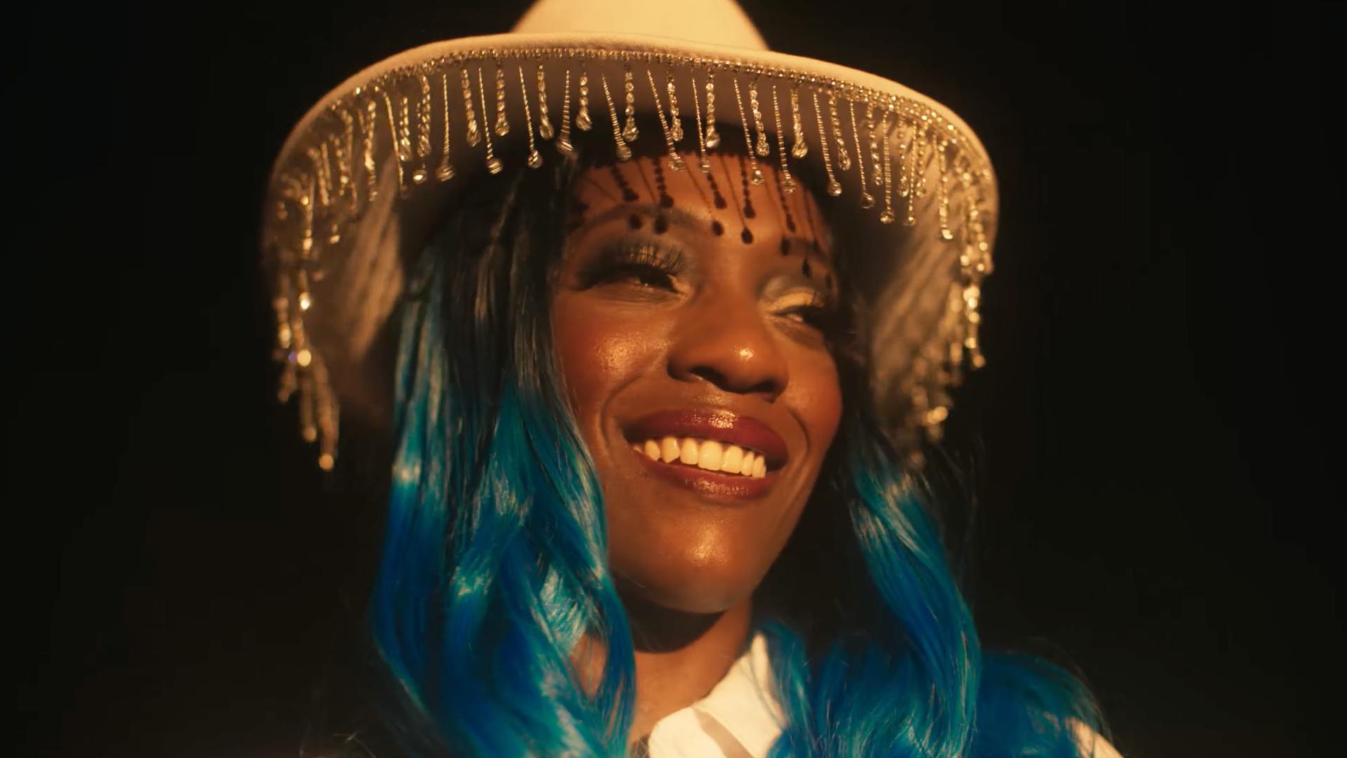 A film still from Ree’s Destiny shows the face of a Black woman who smiles looking into the distance. Her black curled hair fades into a vibrant blue, and she wears a full face of makeup along with a white cowboy hat wrapped with dangling clear crystals around the rim.