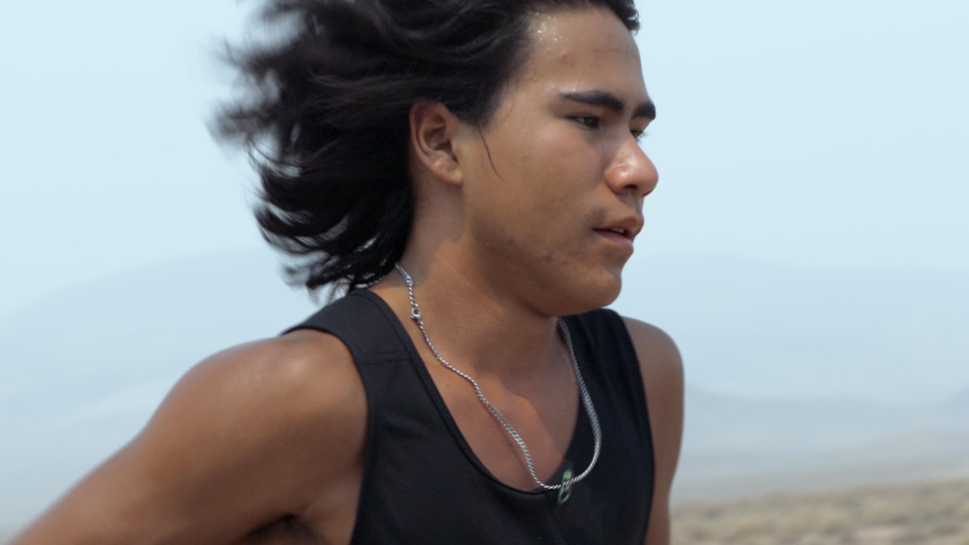 A film still from Remaining Native shows a Native American boy in motion, presumably running, with his mid-length black hair flowing behind him. He wears a black tank top and a chain necklace that holds a soda can tab.