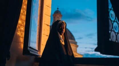 A film still from Run, Sister Joan shows a Black nun standing at an opened window. The side of her face is illuminated by the light from outside as she looks over her shoulder at the camera with a serious face.