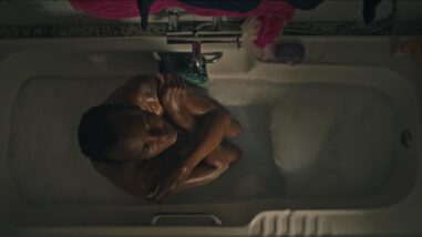A film still from Sabbatical shows an overhead shot of a Black woman curled into a ball in a bathtub. Her arms are crossed in a self-embrace, and she slightly tilts her head upwards with her eyes closed. Her hair is in a low cut.
