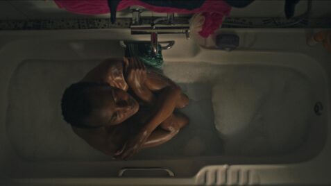 A film still from Sabbatical shows an overhead shot of a Black woman curled into a ball in a bathtub. Her arms are crossed in a self-embrace, and she slightly tilts her head upwards with her eyes closed. Her hair is in a low cut.
