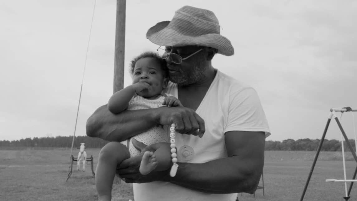 A black-and-white film still from Seeds shows an older Black man holding a baby against his chest. The baby looks out while the man looks down at the baby holding their pacifier. There is a playset in the background.