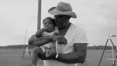 A black-and-white film still from Seeds shows an older Black man holding a baby against his chest. The baby looks out while the man looks down at the baby holding their pacifier. There is a playset in the background.