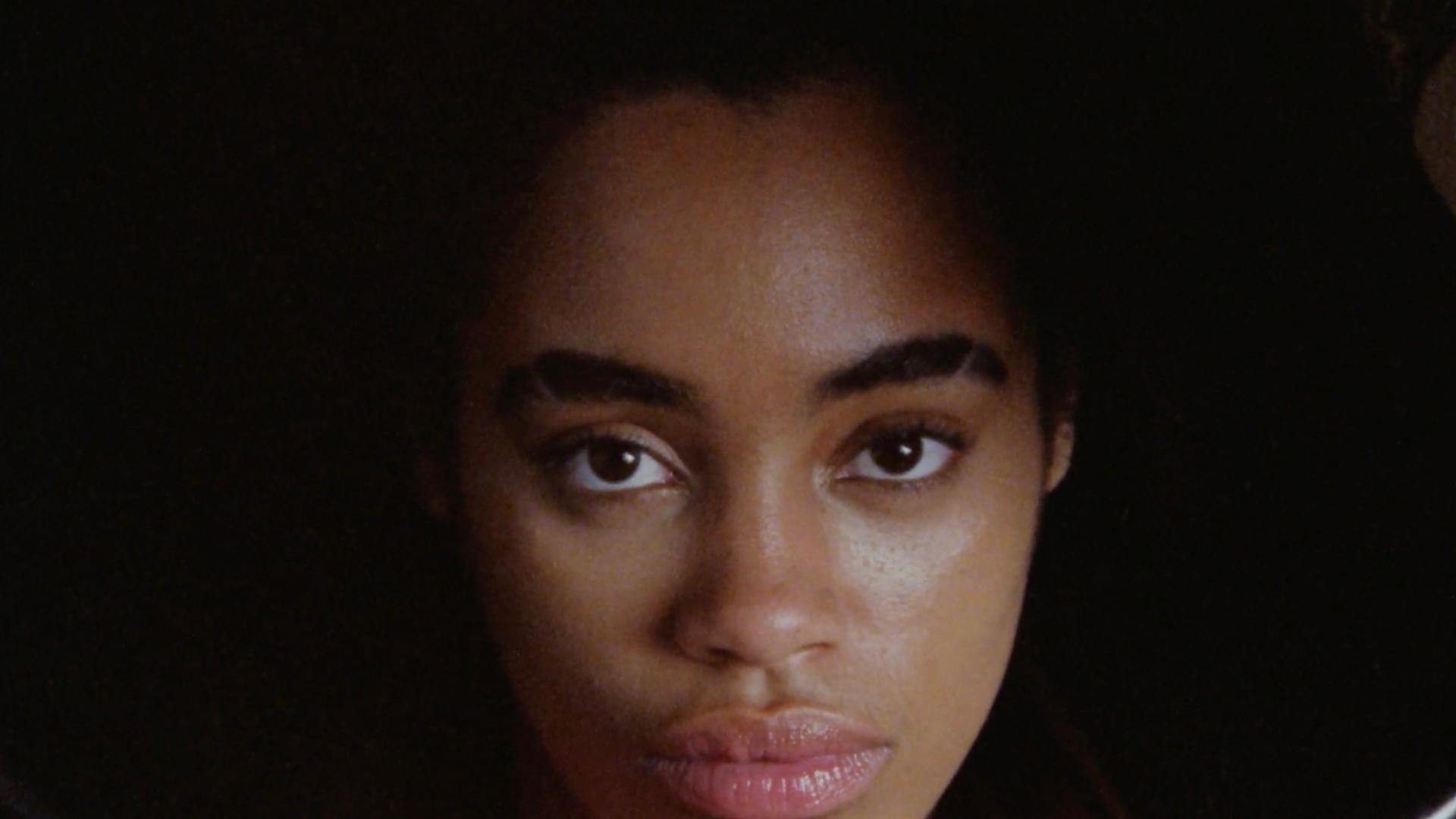 A film still from Seek No Favor shows the face of a Black woman who looks directly into the camera with a straight face.