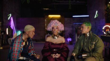 A film still from Space to Breathe shows a Black woman sitting in the middle of two Black men. They are in a room lined with LED strips in various colors. The woman looks into the camera, wearing a purple and pink afro, thick gold necklace, and burgundy off the shoulder dress. The man to her left wears a knitted cap with a kente cloth top and gold jewelry. The other man has two-toned green hair and green jacket along with gold jewelry.