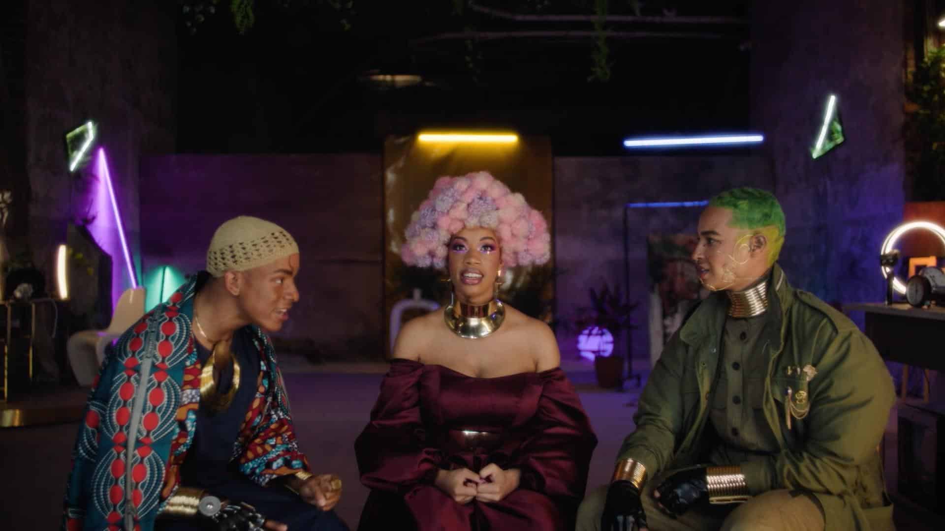 A film still from Space to Breathe shows a Black woman sitting in the middle of two Black men. They are in a room lined with LED strips in various colors. The woman looks into the camera, wearing a purple and pink afro, thick gold necklace, and burgundy off the shoulder dress. The man to her left wears a knitted cap with a kente cloth top and gold jewelry. The other man has two-toned green hair and green jacket along with gold jewelry.