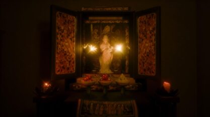 A film still from Spaces as Traces shows an open case in a dark room that holds a figurine of presumably a religious figure. To either side of the figurine are flowers that illuminate the inside of the case. Fruits are placed in bowls directly in front of the open case.