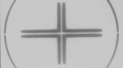 A film still from Speaking in Tongues: Take One shows a white background with a thinly outlined faced black circle. Inside the circle, four black right angled pieces make up a plus sign.