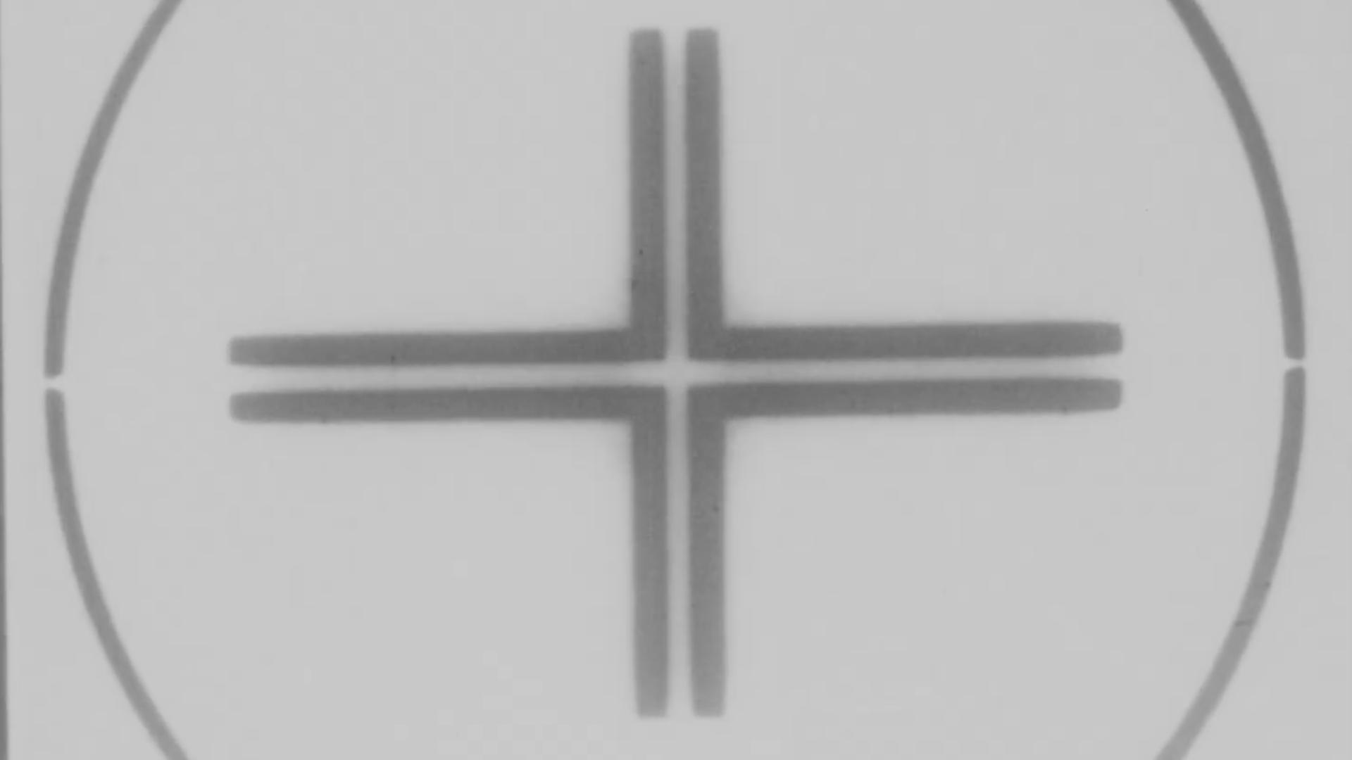A film still from Speaking in Tongues: Take One shows a white background with a thinly outlined faced black circle. Inside the circle, four black right angled pieces make up a plus sign.