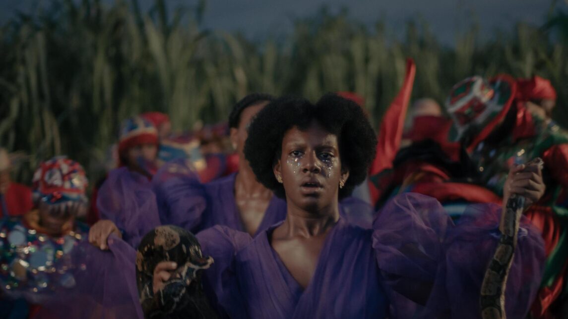 Makenya is wearing a purple dress holding a staff on one hand and a brown snake in the other